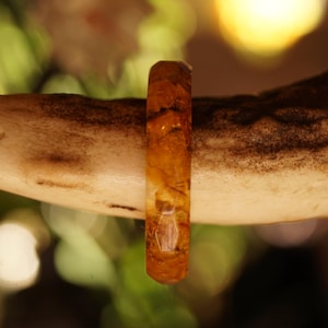 May include: A close-up of a handcrafted ring made of amber-colored resin, showcasing natural inclusions. The ring is set against a blurred background of a light brown antler, highlighting the ring's unique design and color.