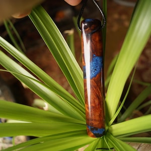 May include: A handcrafted wooden pendant with a rich, reddish-brown hue and striking blue accents. The cylindrical pendant is suspended from a black cord, showcasing a unique blend of natural materials and artistic design. The pendant is approximately 3 inches long.