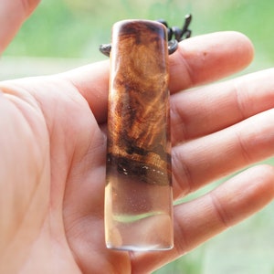 May include: A brown and clear resin pendant with a wood grain pattern. The pendant is hanging from a black cord.