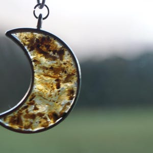 Peut inclure: Pendentif en forme de croissant de lune avec un cadre argenté et un centre texturé de couleur ambre. Le pendentif est suspendu à une chaîne délicate. L'arrière-plan est flou, avec des tons verts et gris.
