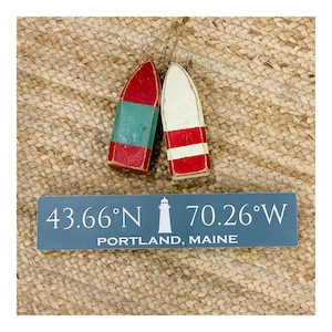 May include: A wooden sign with the coordinates 43.66°N 70.26°W and the text "PORTLAND, MAINE". The sign is painted blue with white lettering and a white lighthouse icon. Two small wooden buoys are in the background.