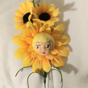 May include: A yellow sunflower brooch with a small porcelain doll face in the center. The doll has blonde hair and painted features.