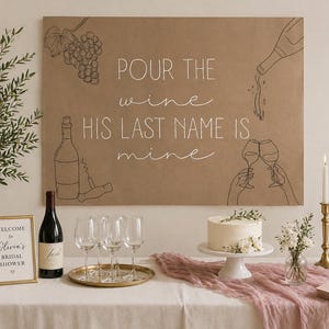 May include: A rectangular brown sign with black line art of grapes, a wine bottle, and two glasses clinking, featuring the white text: POUR THE wine HIS LAST NAME IS mine. The sign is displayed above a table with a cake, wine, and glasses.