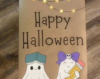 May include: A tan poster featuring the text Happy Halloween in black font. Above the text are yellow string lights. Below are two white ghosts: one wearing a teal book on its head, and the other wearing a purple bow with a skull and holding a pencil.