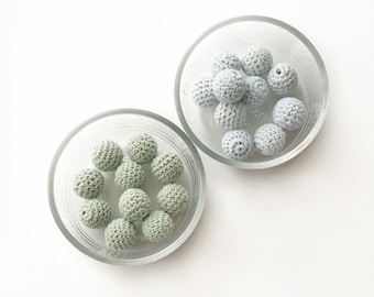 May include: Two glass bowls filled with crocheted beads. One bowl contains light blue beads, and the other bowl contains sage green beads.