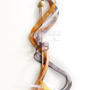 May include: A wooden wand with a round top and colorful ribbons. The ribbons are in shades of gold, silver, white, and black. The wand has the text "MAMA PS PLACE LLC" printed on it.