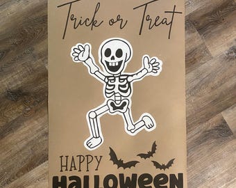 May include: A rectangular tan poster featuring a black skeleton illustration in the center with arms raised. The text Trick or Treat is printed in black at the top, and the text HAPPY HALLOWEEN is printed in black at the bottom with three bats.