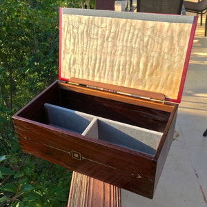 May include: A wooden box with a hinged lid, the lid is open and reveals a light wood interior. The box has a dark wood exterior with a decorative inlay of gold and a gray felt lining.