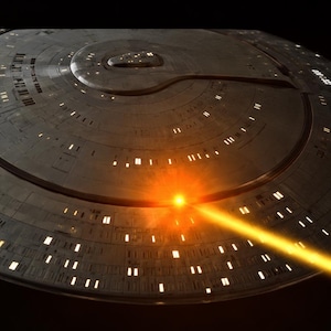 May include: A metallic gray starship saucer section featuring numerous illuminated rectangular windows, a central circular dome, and a bright orange energy beam firing from the hull against a dark black background.