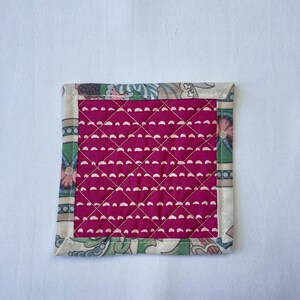 May include: Square quilted coaster with a magenta center and a patterned border. The center features a diamond pattern of white stitches. The border has a floral and leaf design in shades of green, pink, and cream.