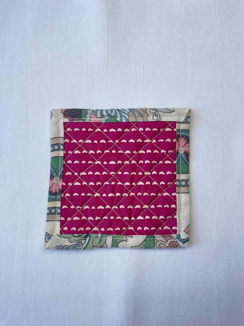 May include: Square quilted coaster with a magenta center and a patterned border. The center features a diamond pattern of white stitches. The border has a floral and leaf design in shades of green, pink, and cream.