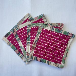 May include: A stack of four square quilted coasters. The coasters have a deep pink center with a white dotted pattern and a floral patterned border in green, pink, and cream. The coasters are arranged on a white surface.