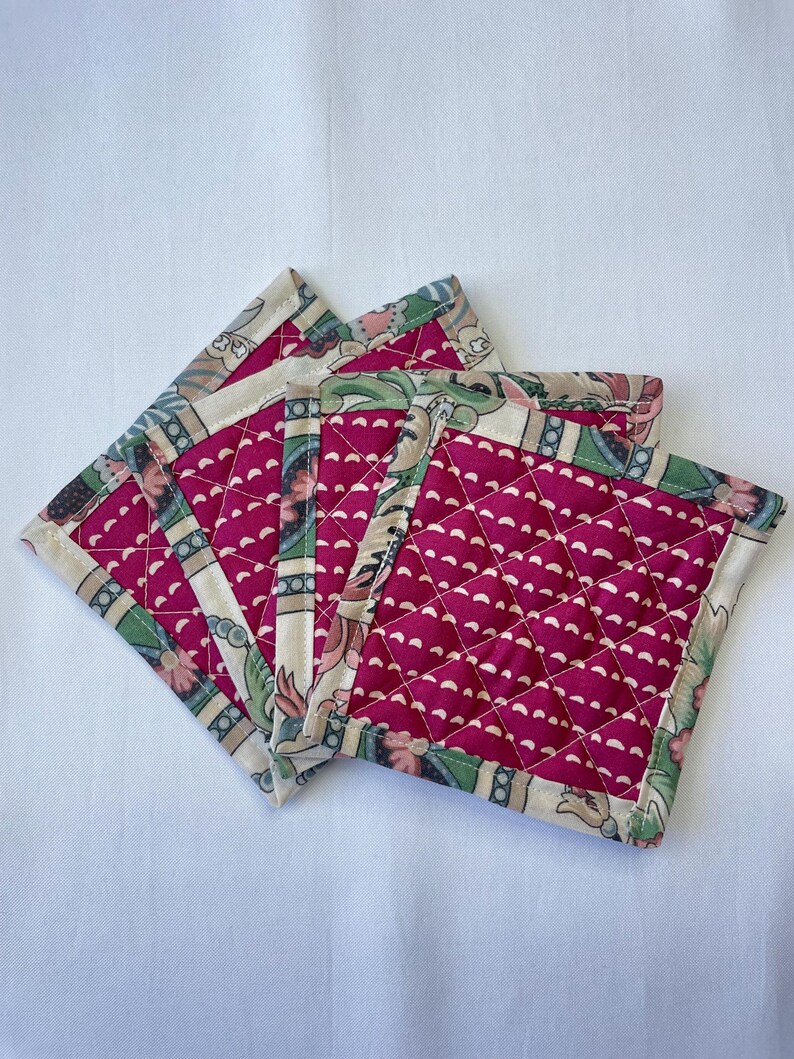 May include: A stack of four square quilted coasters. The coasters have a deep pink center with a white dotted pattern and a floral patterned border in green, pink, and cream. The coasters are arranged on a white surface.