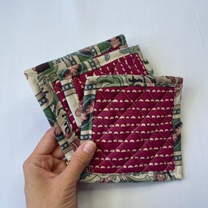May include: A stack of four square quilted coasters. The coasters have a burgundy center with a white dotted pattern and a patterned border with green, beige, and burgundy floral designs. The coasters are held by a hand.