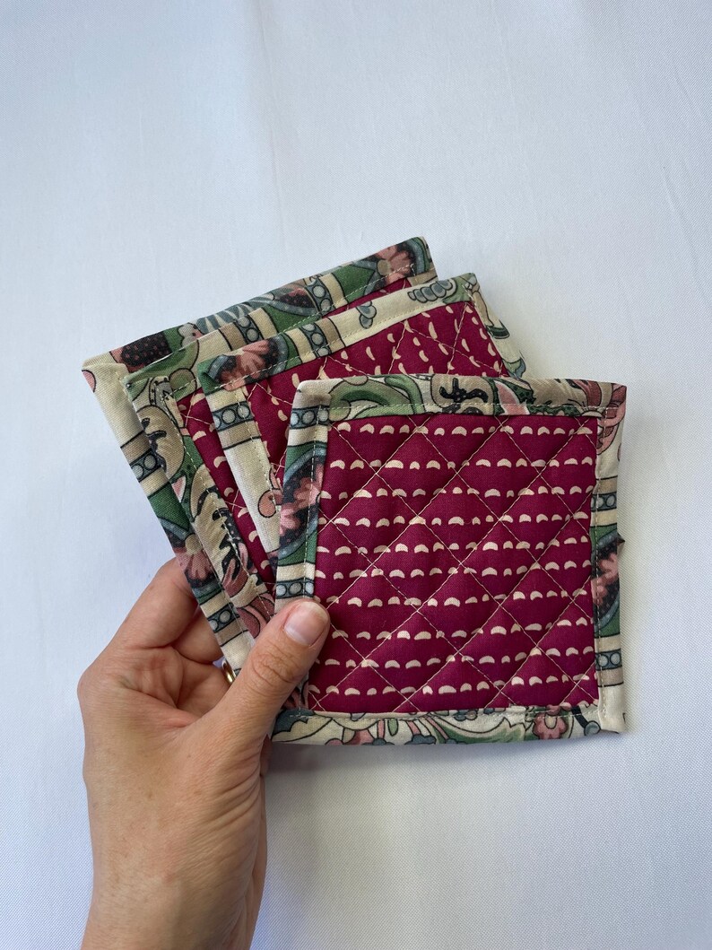 May include: A stack of four square quilted coasters. The coasters have a burgundy center with a white dotted pattern and a patterned border with green, beige, and burgundy floral designs. The coasters are held by a hand.