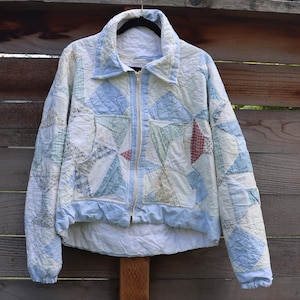 May include: A quilted zip-up jacket featuring a patchwork design in shades of light blue, white, and pale yellow. The garment has a collared neckline, long sleeves with elastic cuffs, and a front zipper closure, constructed from various fabric patterns.