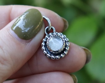 May include: A silver charm with a clear round gemstone set in a beaded border.