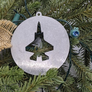 May include: A silver metal Christmas ornament shaped like a circle with a cut-out silhouette of a fighter jet.