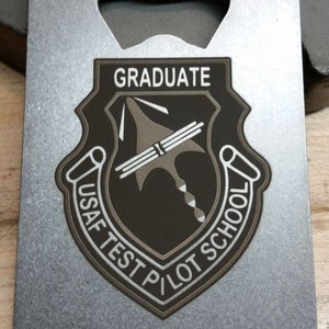 May include: A silver metal bottle opener with a black and silver emblem that reads "Graduate USAF Test Pilot School".