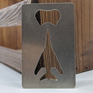 May include: A silver metal bottle opener with a cut-out design of a military aircraft. The opener is sitting on a wooden surface.