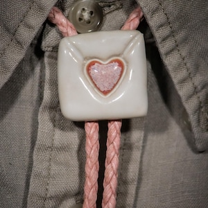 May include: A square, white ceramic bolo tie pendant with a pink heart design. The pendant is attached to a pink braided cord, set against a gray button-down shirt. The heart has a textured appearance.
