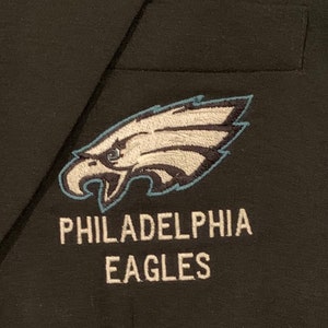 May include: A black blazer with an embroidered Philadelphia Eagles logo. The logo features a white eagle head with a blue beak and a white and blue wing. The text "PHILADELPHIA EAGLES" is embroidered in white below the logo.
