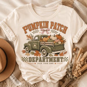 Pode incluir: Uma t-shirt cor de creme com um gráfico de uma carrinha vintage verde carregada de abóboras. O texto diz PUMPKIN PATCH DEPARTMENT PICK YOUR OWN. O design inclui folhas de outono, um laço cor-de-rosa e uma borda axadrezada.