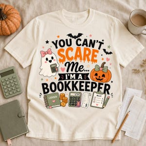 May include: A cream-coloured t-shirt featuring a graphic with the text "YOU CAN'T SCARE ME... I'M A BOOKKEEPER". The design includes a ghost with a pink bow, an orange pumpkin, bats, stars, a ledger, a calculator, coins, and a bills list.