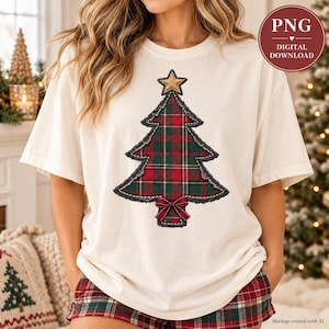 Puede incluir: Una camiseta color crema con un gráfico grande y centrado de un árbol de Navidad con un patrón de cuadros rojos y verdes, una estrella dorada en la parte superior y un pequeño lazo rojo en la base. El diseño tiene bordes deshilachados.
