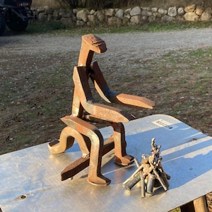 May include: A rustic metal sculpture of a person sitting, crafted from repurposed metal pieces. The sculpture is a warm, rusty brown color and sits on a metal surface. A miniature metal campfire is next to the sculpture.