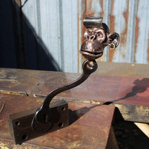 May include: A handcrafted metal sculpture of a monkey's head with a wrench body. The monkey has a bronze finish, a small gold earring, and a stylized metal cap. The sculpture is mounted on a rectangular metal base.