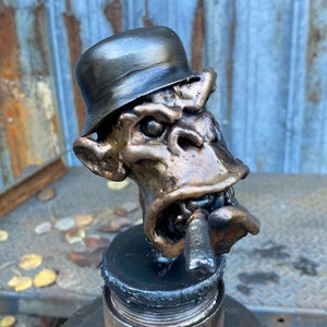 May include: A bronze-colored sculpture of a cartoonish face wearing a hat and smoking a cigar. The sculpture has exaggerated features and is mounted on a metal base. The hat is a dark bronze color, and the face has a distressed finish.