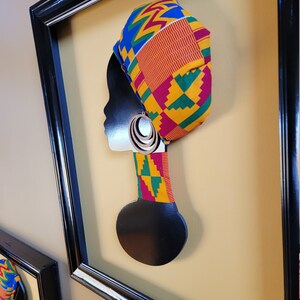 May include: Black silhouette of a person's head with a colorful patterned head wrap and large hoop earrings. The silhouette is mounted on a black wooden base and framed in a black frame.