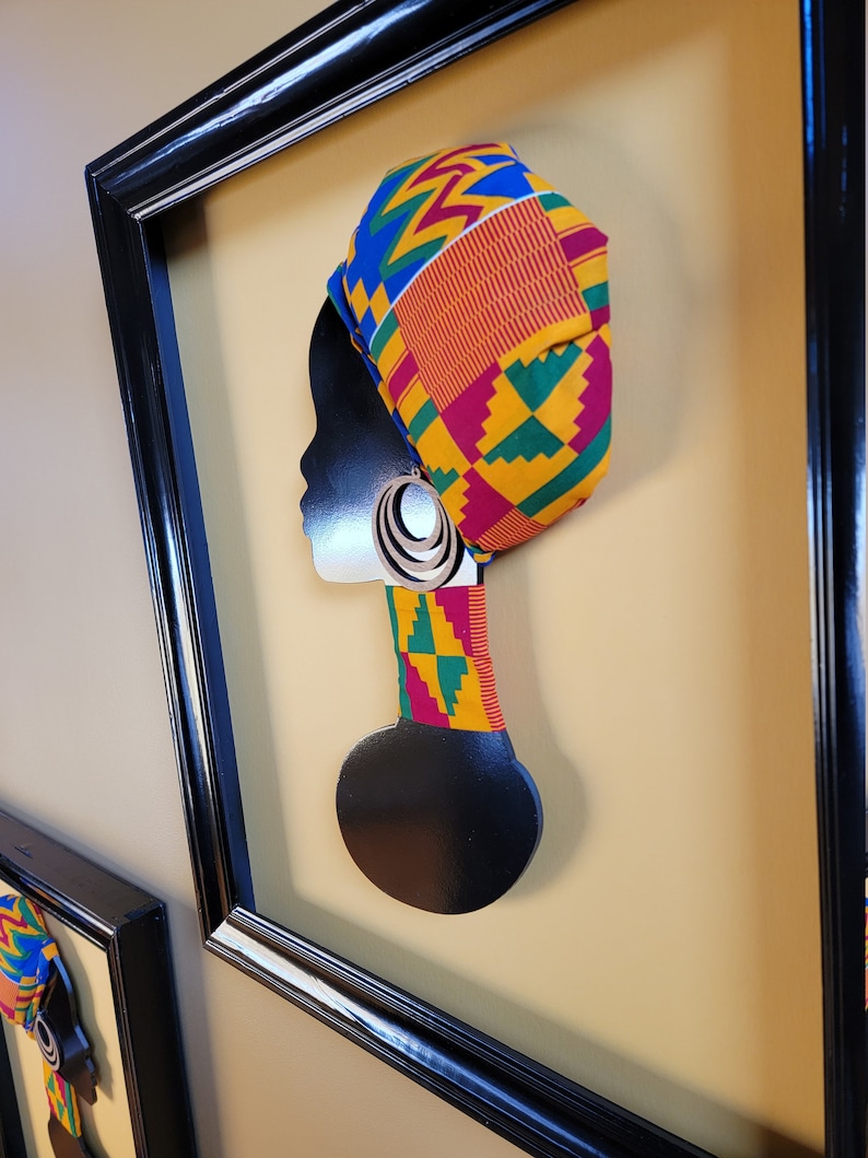 May include: Black silhouette of a person's head with a colorful patterned head wrap and large hoop earrings. The silhouette is mounted on a black wooden base and framed in a black frame.