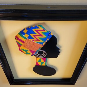 May include: A black silhouette of a woman wearing a colorful patterned head wrap and a matching necklace. The woman is wearing large hoop earrings. The artwork is framed in a black frame.