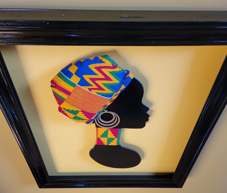 May include: A black silhouette of a woman wearing a colorful patterned head wrap and a matching necklace. The woman is wearing large hoop earrings. The artwork is framed in a black frame.