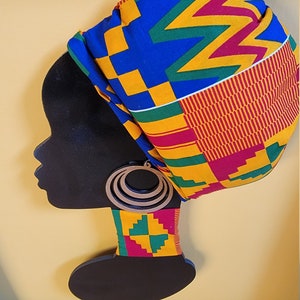 May include: A black silhouette of a woman's head with a colorful patterned fabric head wrap and large wooden earrings. The background is a yellow wall.