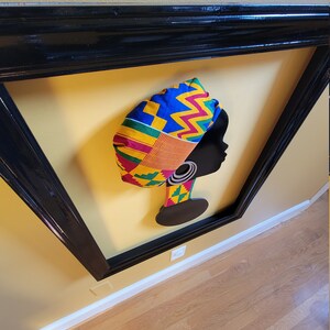 May include: A framed silhouette of a woman wearing a colorful patterned headwrap. The headwrap is made of fabric with yellow, red, green, and blue geometric patterns. The silhouette is black and the background is yellow.