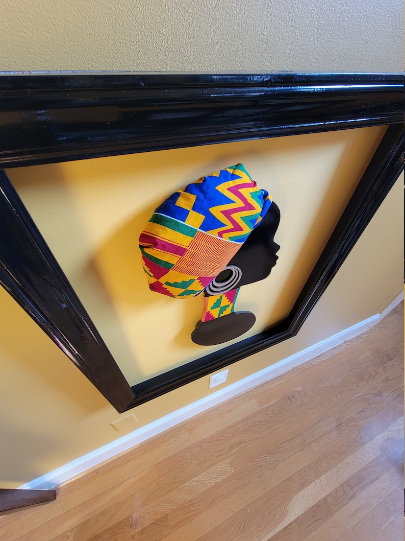 May include: A framed silhouette of a woman wearing a colorful patterned headwrap. The headwrap is made of fabric with yellow, red, green, and blue geometric patterns. The silhouette is black and the background is yellow.