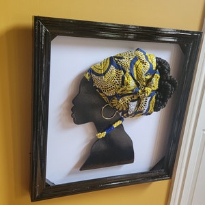 K&ouml;nnte beinhalten: Eine schwarze Silhouette eines Frauenkopfes mit einem gelben und blauen gemusterten Kopftuch, einer Perlenkette und einem goldenen Ohrring. Die Silhouette ist in einem schwarzen Rahmen gerahmt.