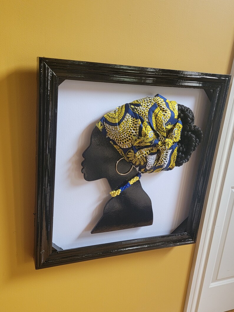 K&ouml;nnte beinhalten: Eine schwarze Silhouette eines Frauenkopfes mit einem gelben und blauen gemusterten Kopftuch, einer Perlenkette und einem goldenen Ohrring. Die Silhouette ist in einem schwarzen Rahmen gerahmt.