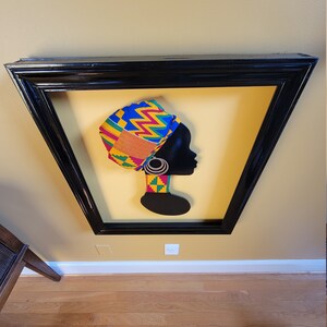 May include: A framed silhouette of a woman wearing a colorful patterned head wrap. The background is a yellow color. The frame is black.