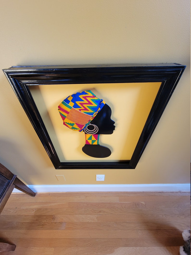 May include: A framed silhouette of a woman wearing a colorful patterned head wrap. The background is a yellow color. The frame is black.