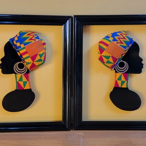 May include: Two framed black and gold silhouette portraits of women wearing colorful head wraps and necklaces. The portraits are set against a yellow background.