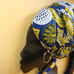 K&ouml;nnte beinhalten: Eine schwarze Silhouette eines Personenkopfes mit einem blauen und gelben gemusterten Kopftuch. Das Kopftuch hat einen goldenen Ohrring und eine blaue und gelbe Perlenkette.