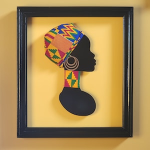 May include: A framed silhouette of a woman's head with a colorful patterned head wrap and large earrings. The background is a light yellow.