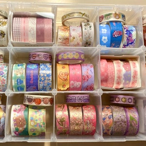 Puede incluir: Una caja de almacenamiento de pl&aacute;stico transparente llena de rollos de cinta washi de colores. La cinta washi est&aacute; organizada en filas y columnas, con cada rollo teniendo un patr&oacute;n diferente. Los patrones incluyen flores, animales, formas geom&eacute;tricas y m&aacute;s.