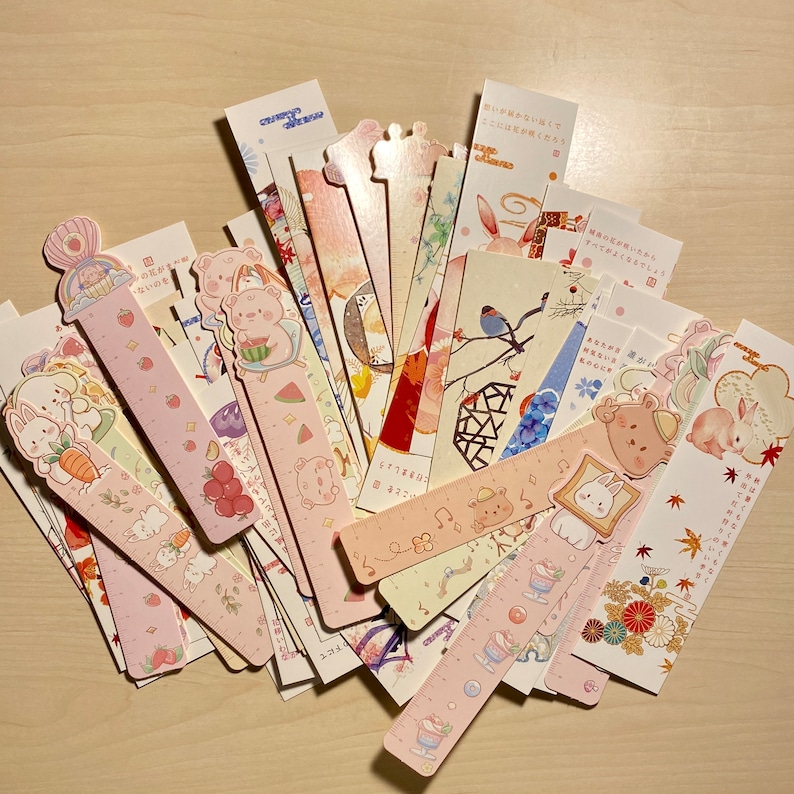 May include: A collection of pink and white bookmarks with various illustrations, including animals, flowers, and geometric patterns. Some bookmarks feature Japanese text, such as "すべてがよくなるでしょう" which translates to "Everything will be fine".