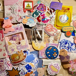 Puede incluir: Una bolsa de pl&aacute;stico transparente llena de pegatinas de colores. Las pegatinas presentan animales de dibujos animados, flores, comida y otros dise&ntilde;os. La bolsa tambi&eacute;n incluye algunas tarjetas peque&ntilde;as con texto, como "Hey! Have a nice day!" y "I'm sorry."