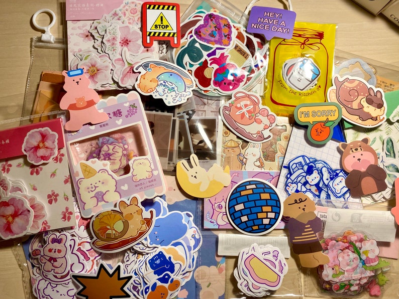 Puede incluir: Una bolsa de pl&aacute;stico transparente llena de pegatinas de colores. Las pegatinas presentan animales de dibujos animados, flores, comida y otros dise&ntilde;os. La bolsa tambi&eacute;n incluye algunas tarjetas peque&ntilde;as con texto, como "Hey! Have a nice day!" y "I'm sorry."